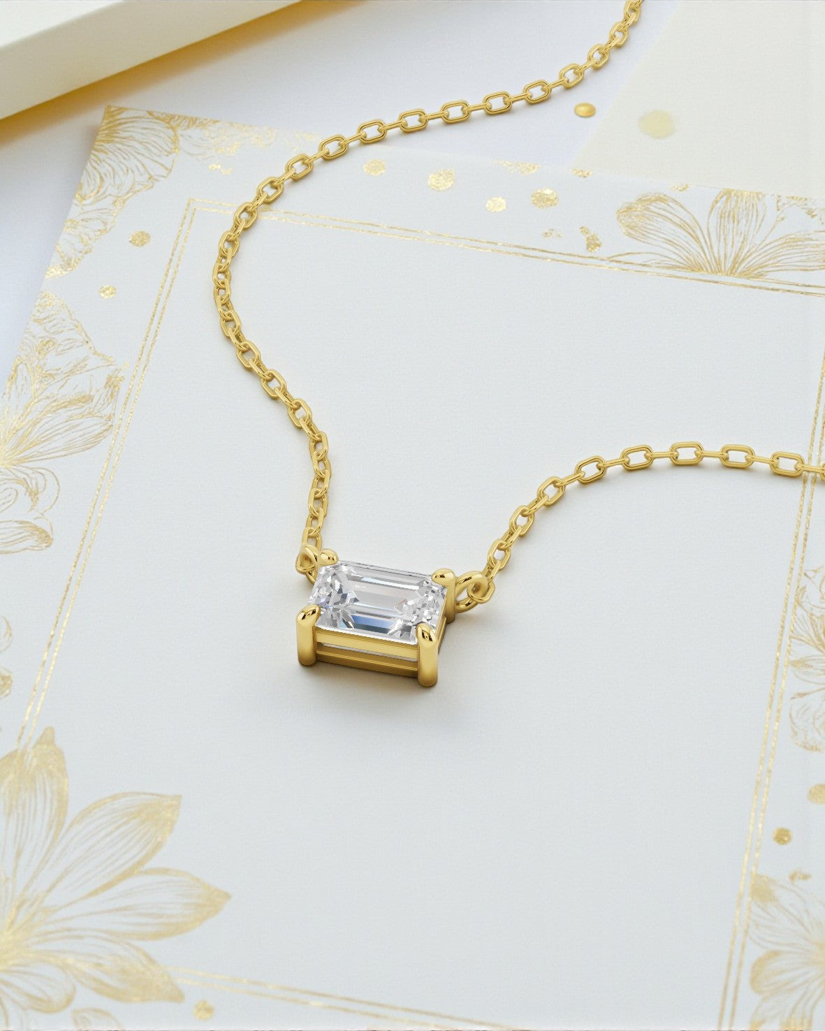 エメラルドカット（長方形）のダイヤモンドを水平にセットした、イエローゴールドのネックレス。金色の花の線画が描かれた白いカードの上に置かれています。