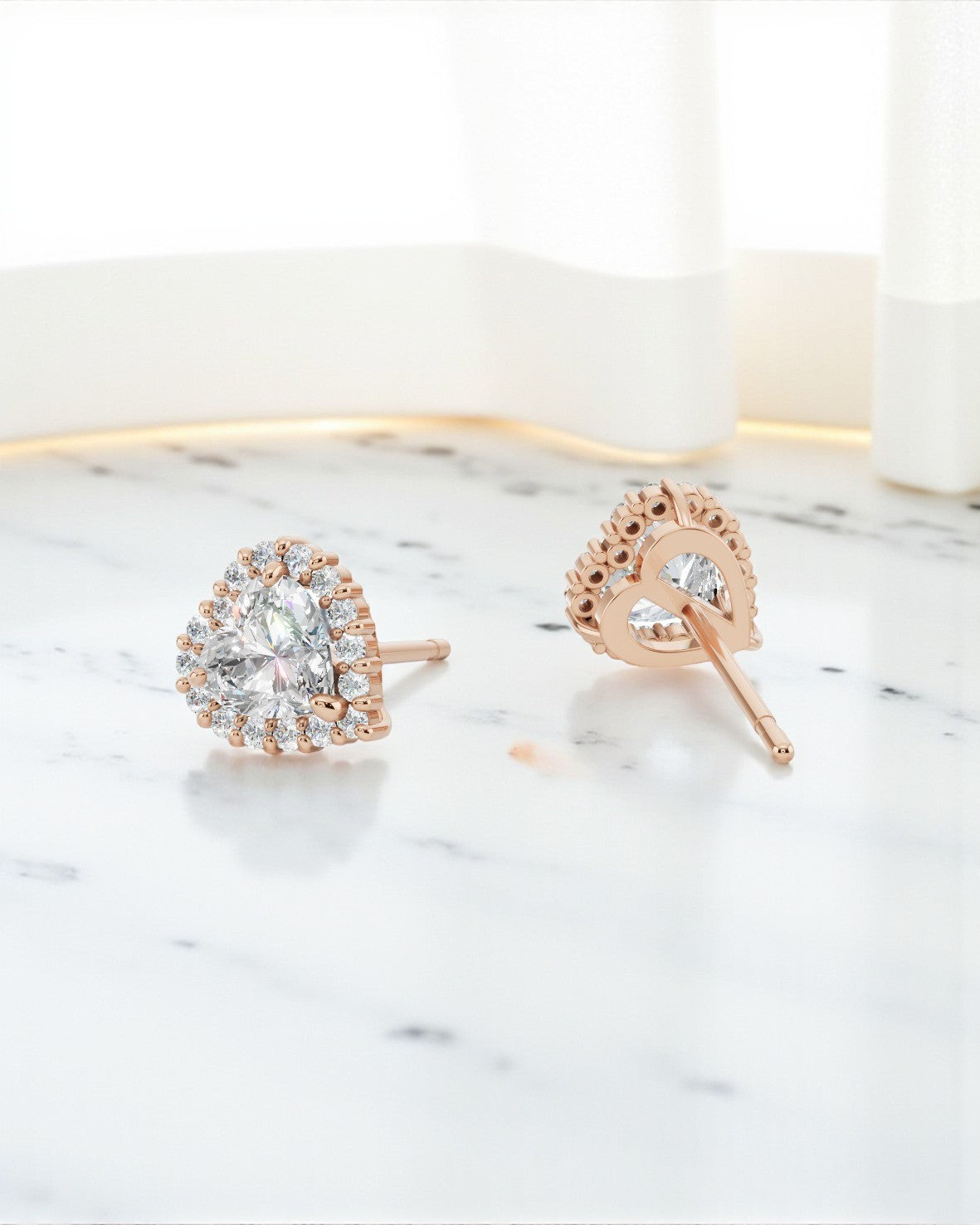 ハートシェイプのダイヤモンドをヘイローセッティングで囲んだ、ローズゴールドのピアス一組。白い大理石の表面に置かれており、片方はポスト（留め具の軸）が見えるように横向きになっています。