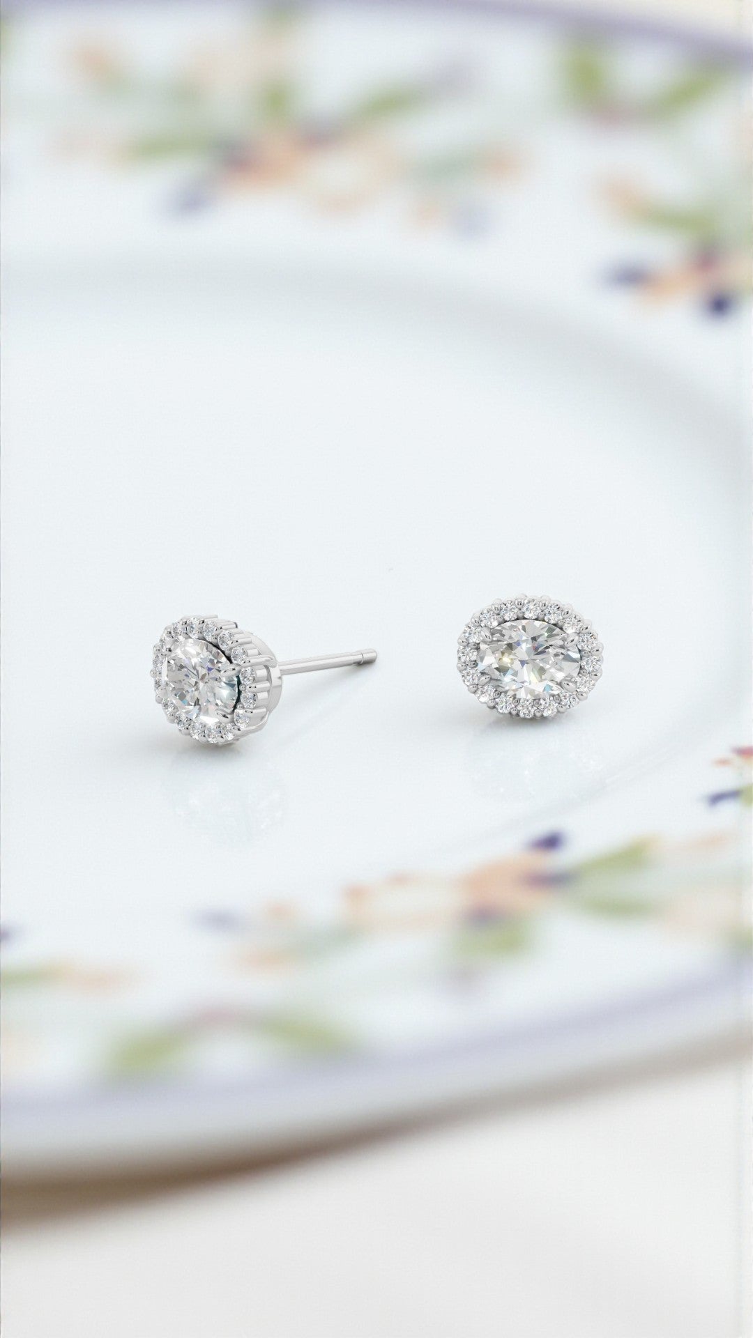 オーバルカット（楕円形）ダイヤモンドホのワイトゴールド（またはプラチナ）のヘイローピアス一組。淡い花柄が描かれた白い磁器の皿の上に置かれています。