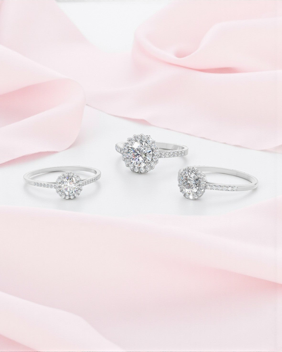3つのラウンドヘイローダイヤモンドリングが、ピンク色のサテン生地がドレープする白い背景に配置。すべてホワイトゴールド（またはプラチナ）製で、中央にラウンドカットのダイヤモンドがセット。