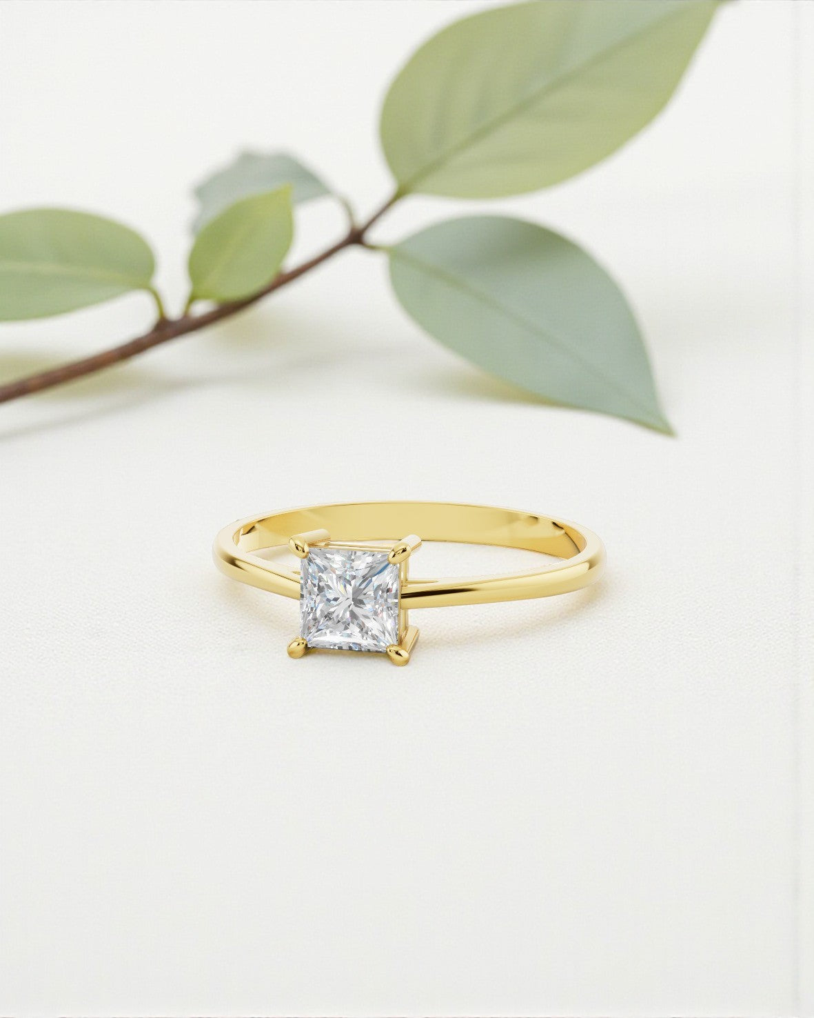 プレーンなイエローゴールドのバンドに、プリンセスカット（四角形）のダイヤモンドを4本爪で留めたソリティアリング。背景には、ぼかした緑の葉が映る白い平面が広がっています。